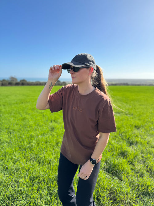 A woman wearing a brown t-shirt, black pants, a black cap, and sunglasses stands in a grassy field under a clear blue sky.