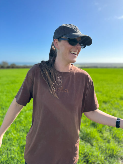 A woman wearing a brown t-shirt with a subtle 'ella' logo, a dark cap, and sunglasses, standing in a green field under a clear blue sky.