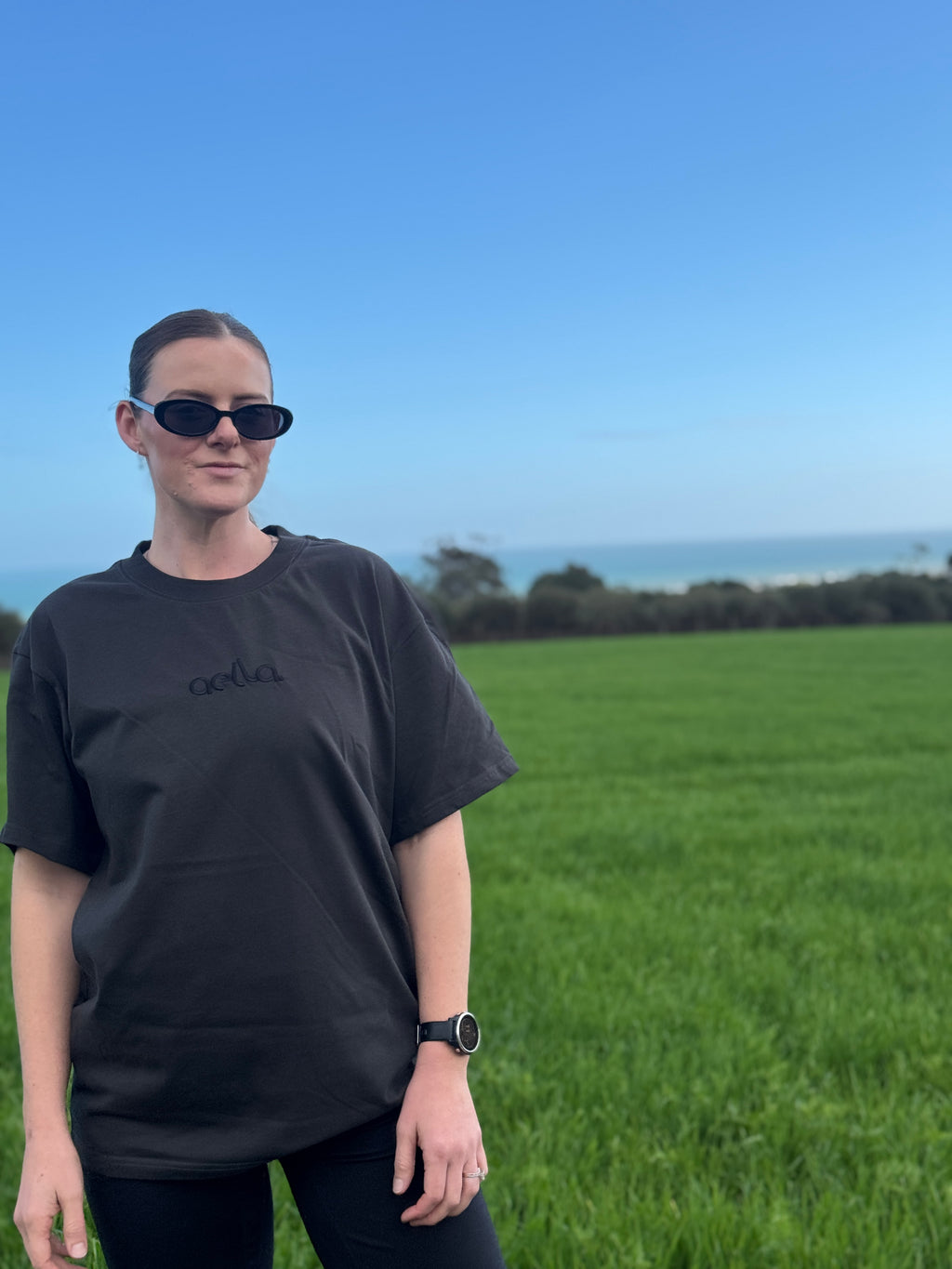 A woman wearing a black oversized t-shirt with a subtle tonal Aella logo, black sunglasses, and a watch, standing in a green grassy field with the ocean and blue sky in the background.