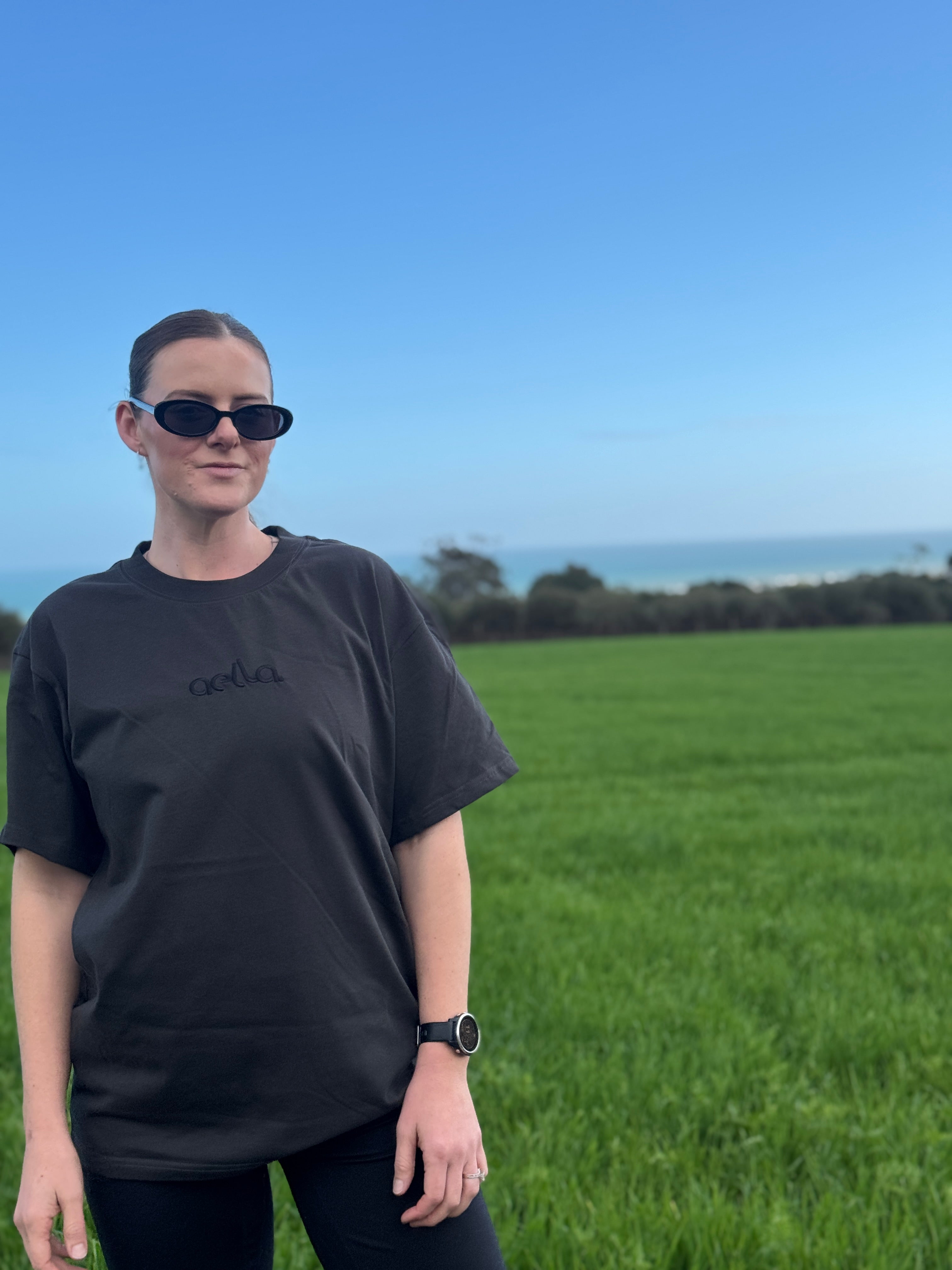 A woman wearing a black oversized t-shirt with a subtle tonal Aella logo, black sunglasses, and a watch, standing in a green grassy field with the ocean and blue sky in the background.