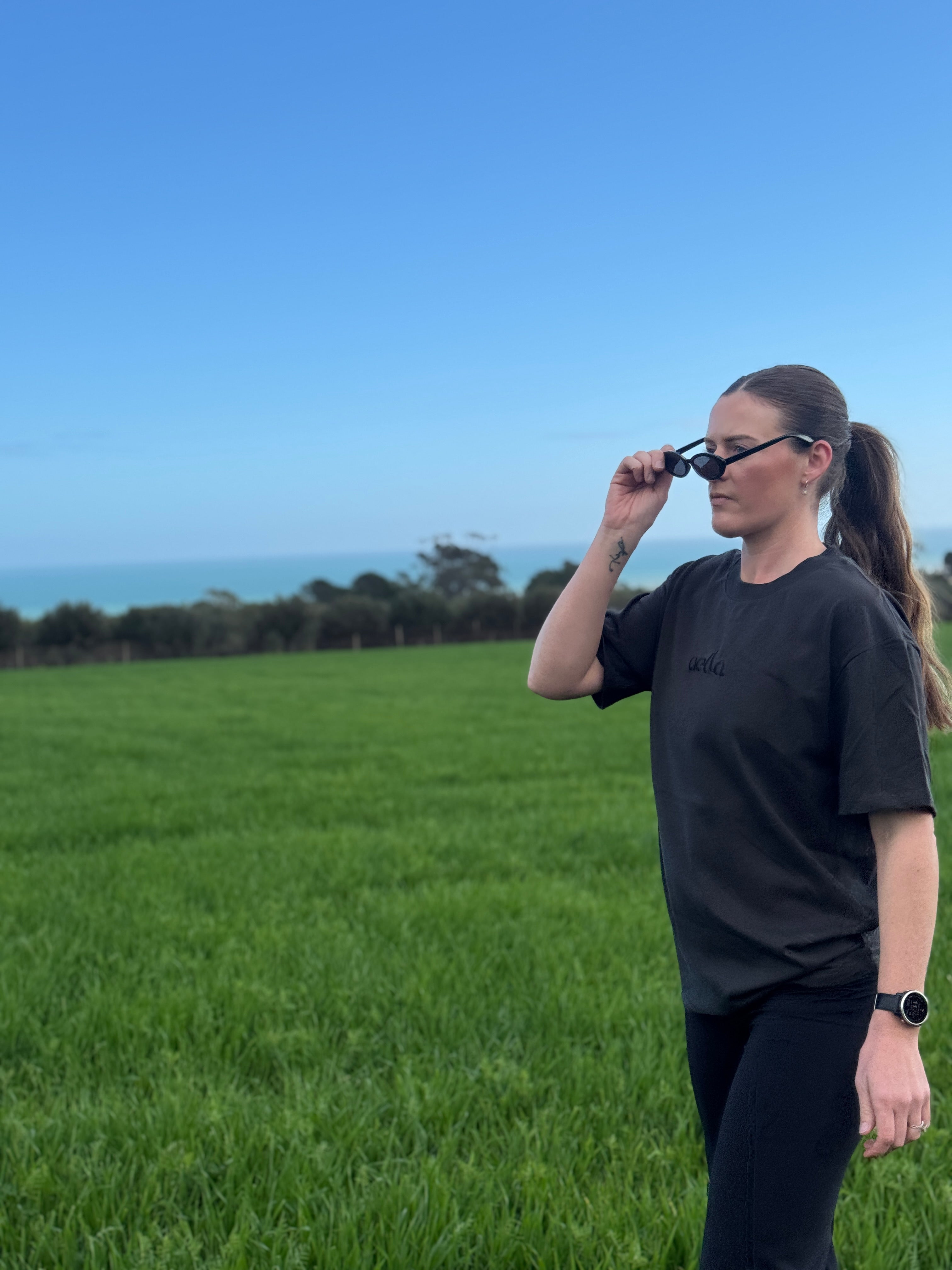 A woman in a black t-shirt and black pants stands in a grassy field outdoors, holding her black sunglasses to her face. The background features a green field, a line of trees, and a clear blue sky.
